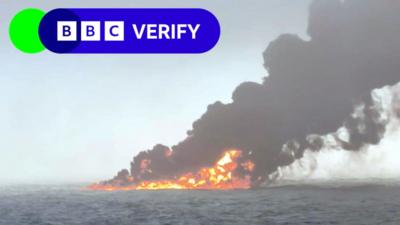 A grainy photo taken in the north sea off the east coast of England. It shows oil on the surface of the water that is on fire, with bright orange flames and a huge plume of dark grey smoke billowing into the sky. 