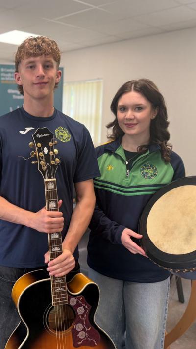 Two teenagers, a male and female, both with long brown hair. Jacob wears a navy uniform tshirt and is holding a guitar. Niamh is also in uniform, a green and navy half zip, holding a bordhan. They are pictured beside grey chairs and a green Glengormley School of Traditional Music backdrop.