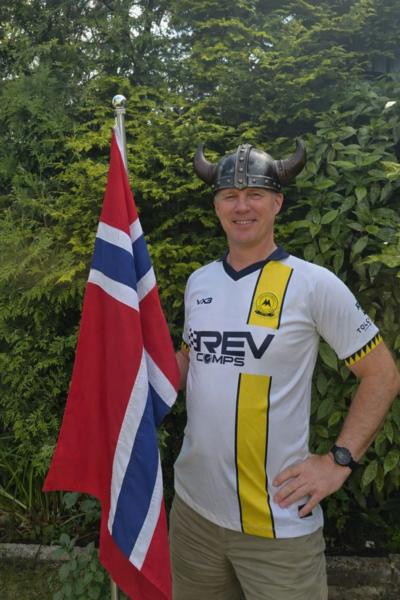 Man in a white and yellow Torquay United football top wears a Viking helmet and holds a red and blue and white Norweigan flag in front of a green hedge 