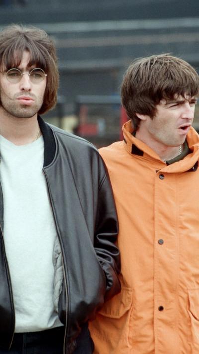 Two people stand side by side outdoors. The person on the left wears round sunglasses, a black leather jacket, and a pale shirt. The person on the right wears a bright orange jacket with a high collar and buttons.