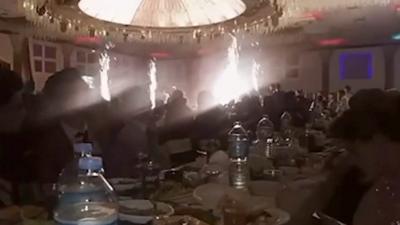 Long table full of people and food, as sparklers go off in the background