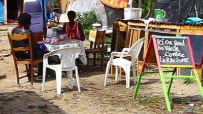 Mauritanian man sitting at a table with BBC reporter, sign in front reads, in French, 'Vaccines against racism on sale here'