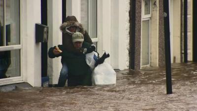 Man giving a woman a piggyback through thigh-deep flood water