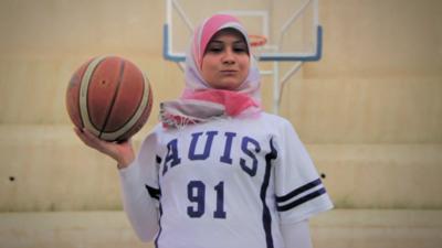 Woman wearing headscarf, sports clothing and holding a basketball.