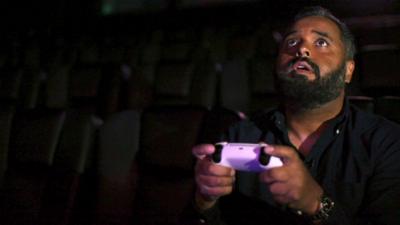 Click reporter Marc Cieslak holds a gaming controller while playing a game on a cinema screen