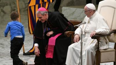 A boy coming from the audience onto the stage runs past Pope Francis