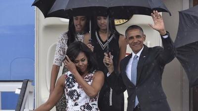 President Barack Obama, Michelle Obama and daughters walking down stairs from a plane. All holding up large black umbrellas.