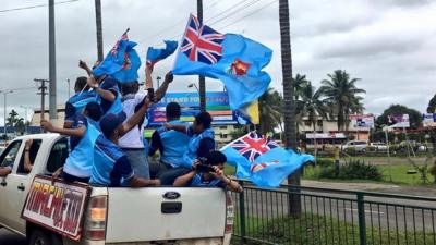 Men in the back of a pickup truck in Fiji T-shirts and waving Fiji flags