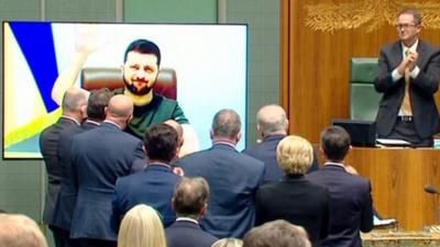 Australian parliament standing up and clapping at a TV screen with President Zelensky on