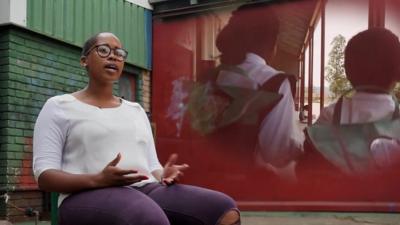 Thato Kgatlhanye sits in front of a video projection showing two children wearing her solar backpacks.