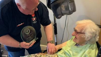 David Hardy bending down to show an elderly woman in a lime green top his glass Volunteer of the Year award