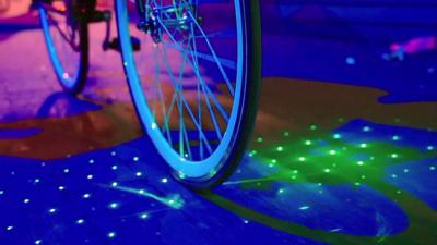 A bicycle being ridden in the dark, green dots surround the front wheel are on the road