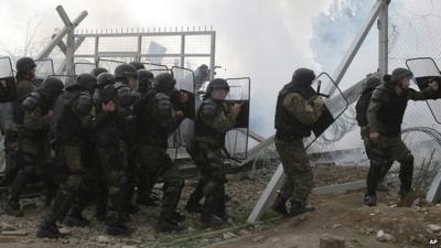 Stranded refugees and migrants break an iron fence and throw stones from the Greek side of the border as Macedonian policemen use tear gas,