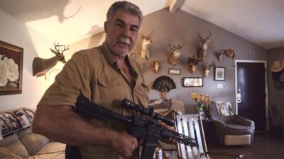 Man holding AR-15 in front of a wall hung with deer heads