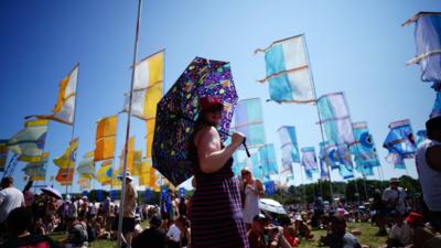 A woman using an umbrella for shade stands in a field in front of festival-goers, flags and a blue sky