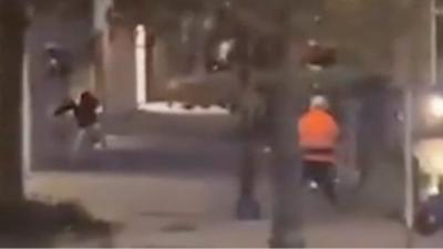 Man in orange shirt aims gun at people in a dark street