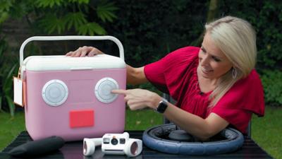 Lara Lewington points at a pink cool box which has speakers on the front. Other gadgets are on the table