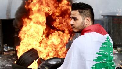 Person with Lebanese flag in front of burning tyres, 15 July 2021