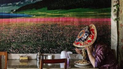 A lady in a hat eats noodles next to a large picture of a landscape