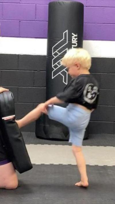 A small child with blonde hair wearing a black T-shirt and shorts is kicking a boxing pad.  Behind her is a black punch bag next to a wall.