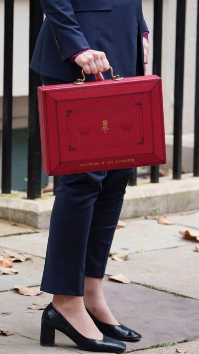 The bottom half of a woman, the chancellor, holding a red briefcase wearing a navy suit and block heels