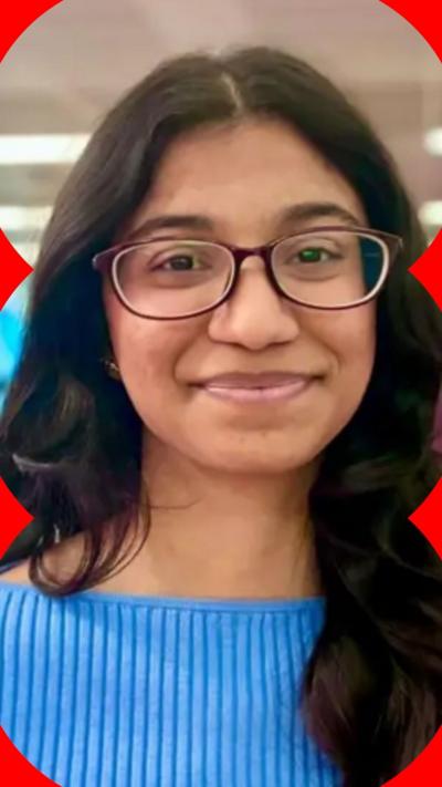 A woman wearing a blue top stands and smiles at the camera. She wears glasses and has long dark hair.