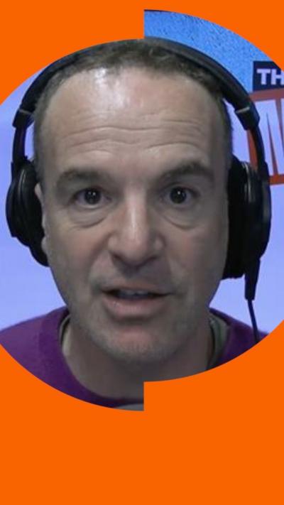 A man, Martin Lewis, has short brown hair and wears a purple jumper. He sits in front of a microphone and is wearing headphones.