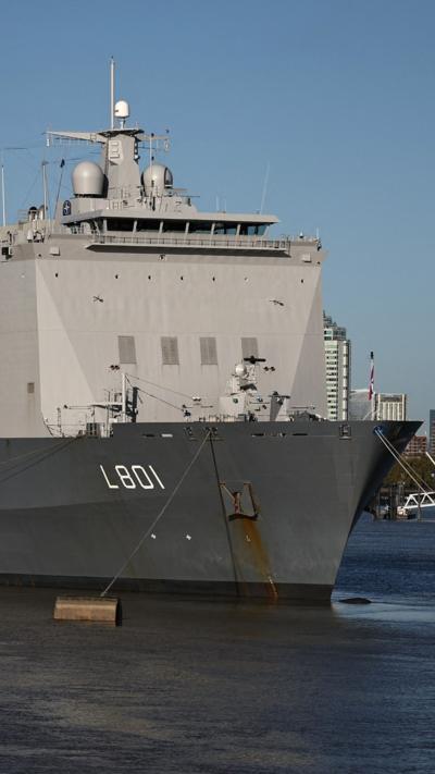 A large naval vessel, anchored in the River Thames with a city skyline in the background, under a clear blue sky.