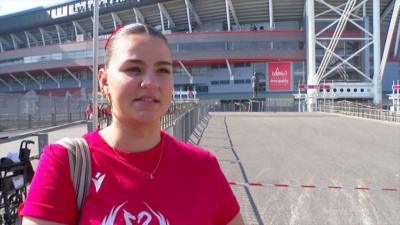 Rugby fan outside of the Principality stadium, she is wearing a red T-shirt and has her dark hair scraped back into a bun. She is also wearing gold hoop earrings