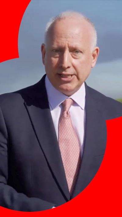 Steve Rosenberg looks directly at the camera. He stands in-front of a plume of smoke in the background. He wears a navy suit and pink tie.