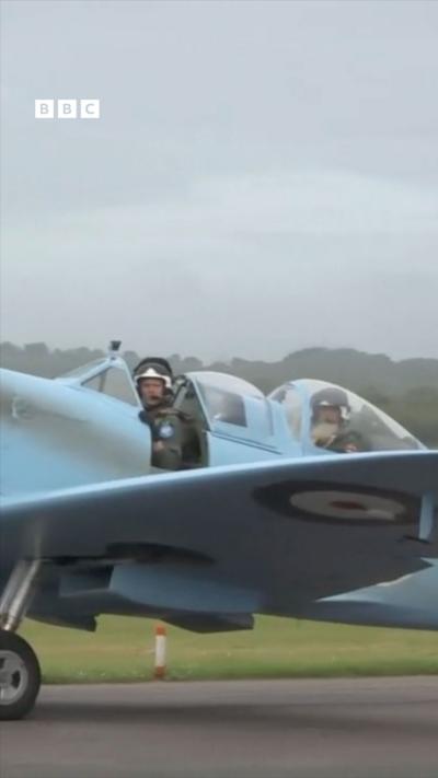 A man in a cockpit of a Spitfire on a runway, the sky is covered in grey cloud