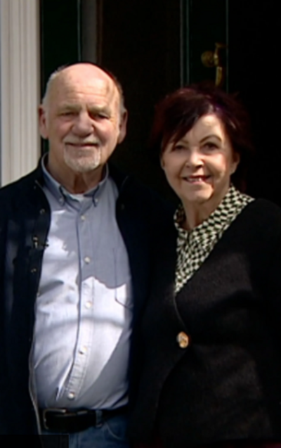 a couple stood in front of a black door. man has a blad head, blue shirt and dark blue coat. the woman on right had red hair, chequered shirt and black cardigan.