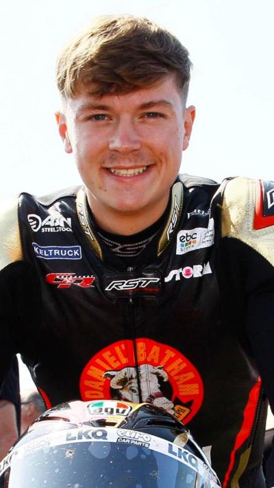 A young man with brown hair in a black motorcycle outfit with white shoulders and an orange circle in the middle