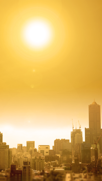 A panoramic view of a city with the sun directly overhead, casting an orange glow across the sky from the intense heat