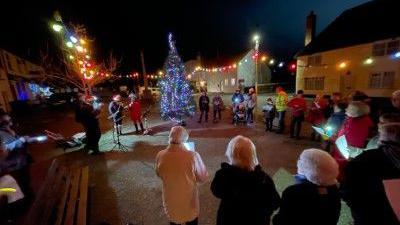 People standing in a circle around a lit up Christmas tree. They are singing carols.