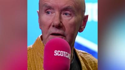 A man wearing a yellow shirt speaks into a pink microphone