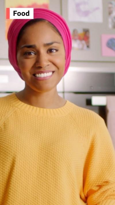 A woman, chef Nadiya Hussain, wears a yellow jumper and a pink head scarf and smiles