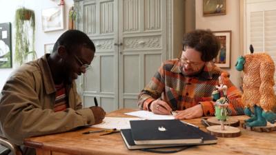 De-Graft and Mikey Please are sat down at a table and drawing