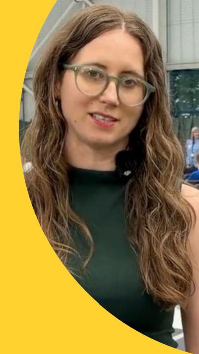 A woman with glasses and long brown hair talks to the camera