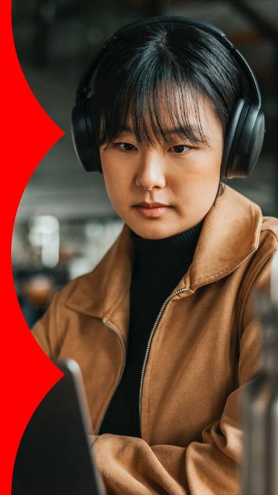 A woman in an office wears wireless headphones and looks at a laptop 