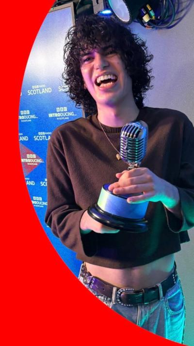Jacob Alon, smiling, holding a trophy, wearing a cropped brown jumper and jeans.