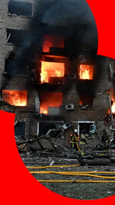 An emergency worker with a hose stands in front of a damaged and burning building with fire visible in multiple, blown-out windows. Thick smoke rises into the air.