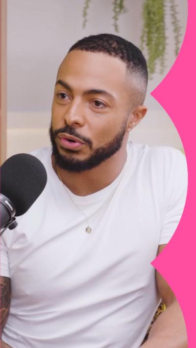 Tyler West sits in a white t-shirt with a black mic in front of him. A hanging plant is behind him.