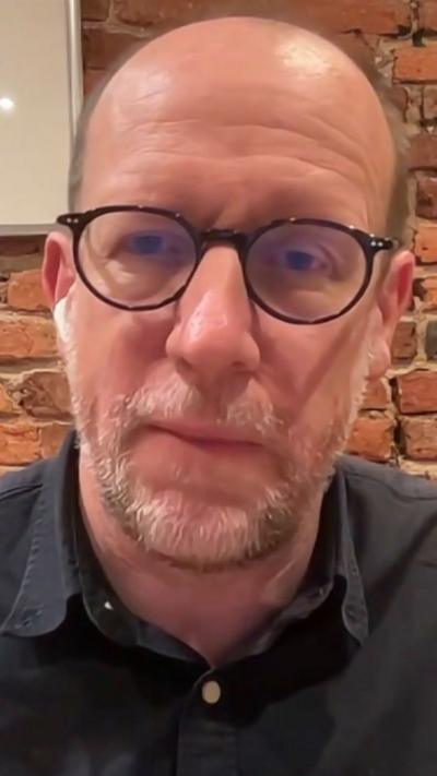 A man in glasses speaks during a virtual interview with a brick wall behind him.