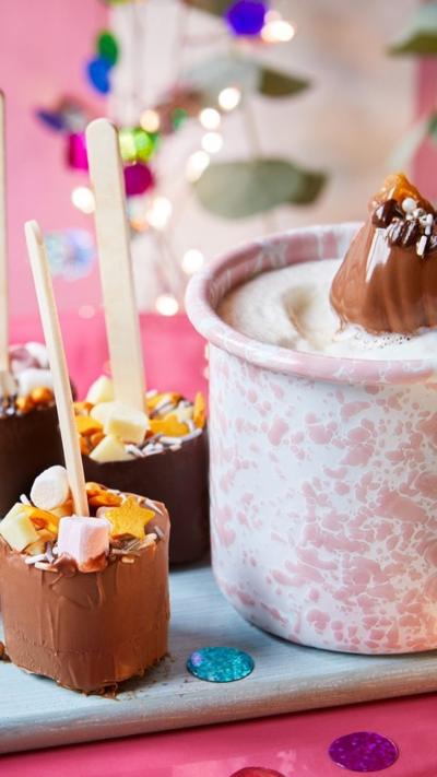 A pink and white mug of hot chocolate with two decorated marshmallows on wooden sticks 