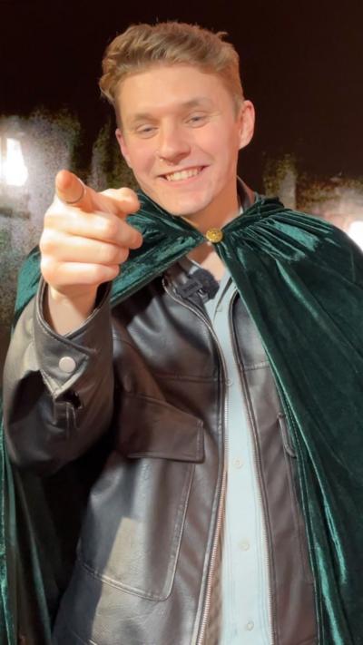 Reece Ward wearing a green traitors cloak points off camera while smiling.