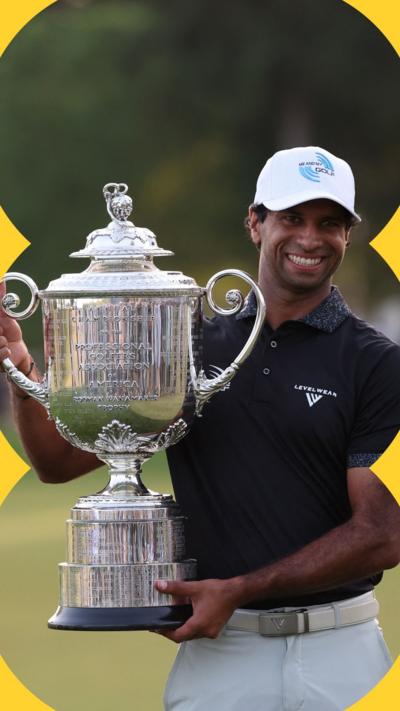 A man wearing a white cap, black polo shirt and white trousers smiles as a he holds a large trophy