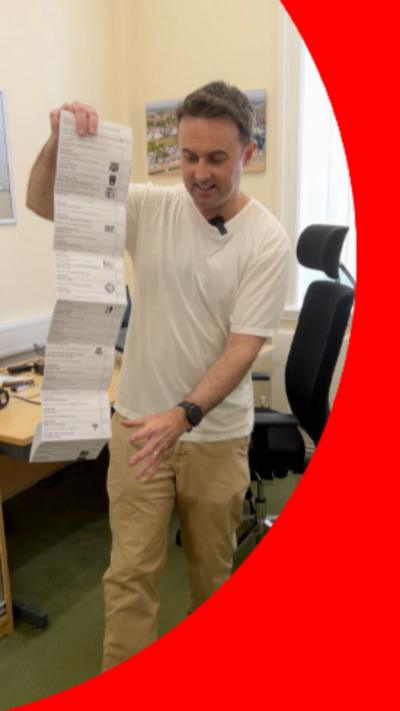 Man with short hair holds a long piece of paper which stretches down by his right side.