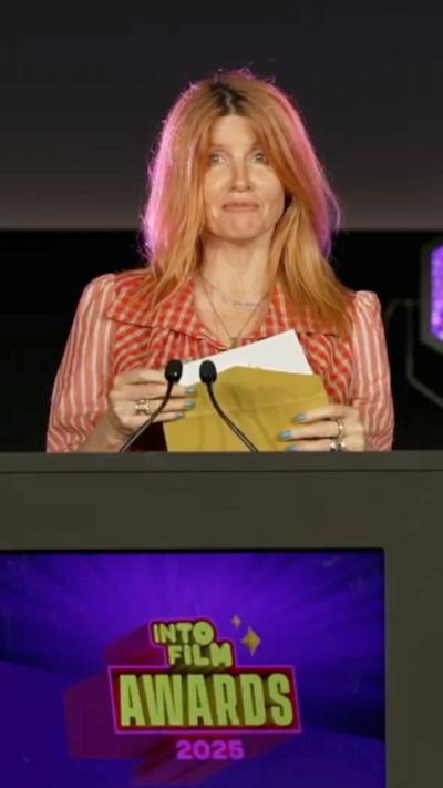 A woman stands behind a podium that says 'Into Film Awards 2025'. She is opening an award envelope. She has shoulder-length blonde hair and is wearing a red and white striped top. 
