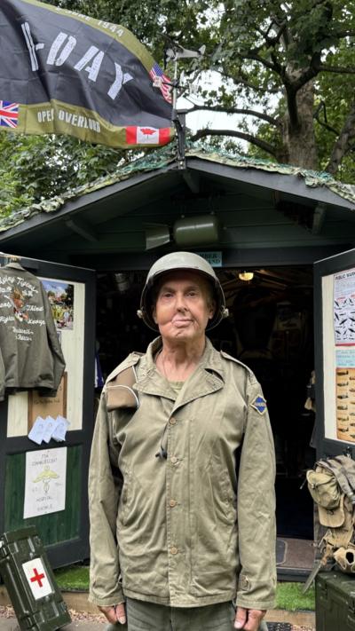 Man in full militry regalia wearing a helmet. Behind him a shed dedicated to WWII memorabilia.